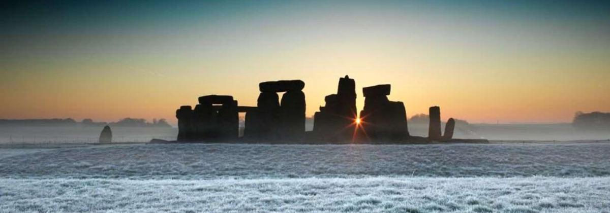 a landscape image of the Stonehenge monument during sunset, with snow on the ground