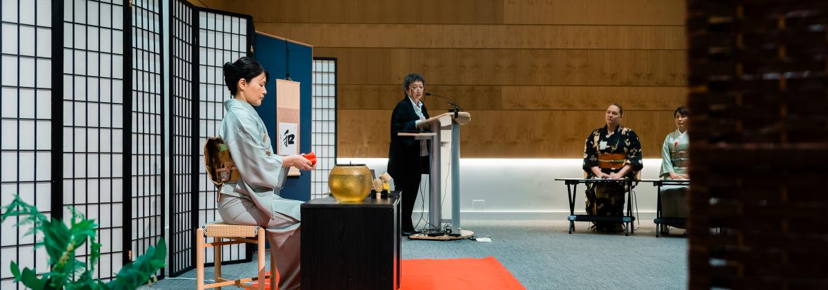  A woman in a kimono is seated at a small black table. Another woman stands at a podium and addresses a crowd.