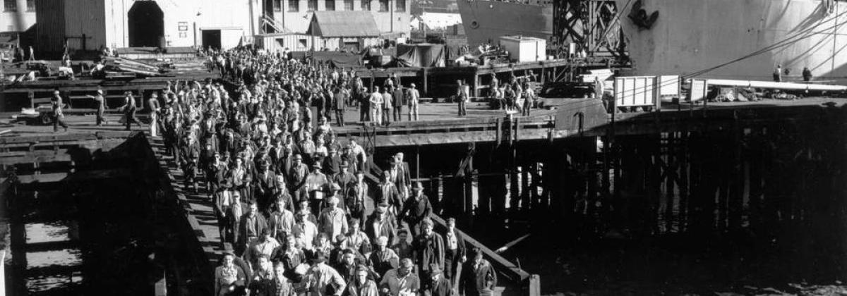 Black and white photo of a large group of workers leaving a ship, walking on a dock. 