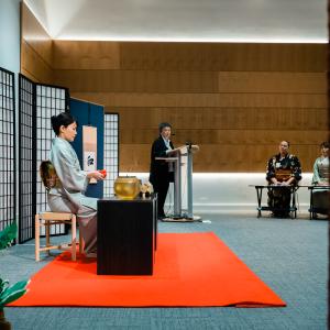  A woman in a kimono is seated at a small black table. Another woman stands at a podium and addresses a crowd.