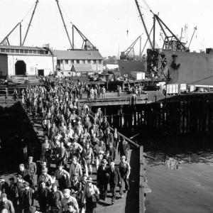 Black and white photo of a large group of workers leaving a ship, walking on a dock. 