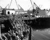 Black and white photo of a large group of workers leaving a ship, walking on a dock. 